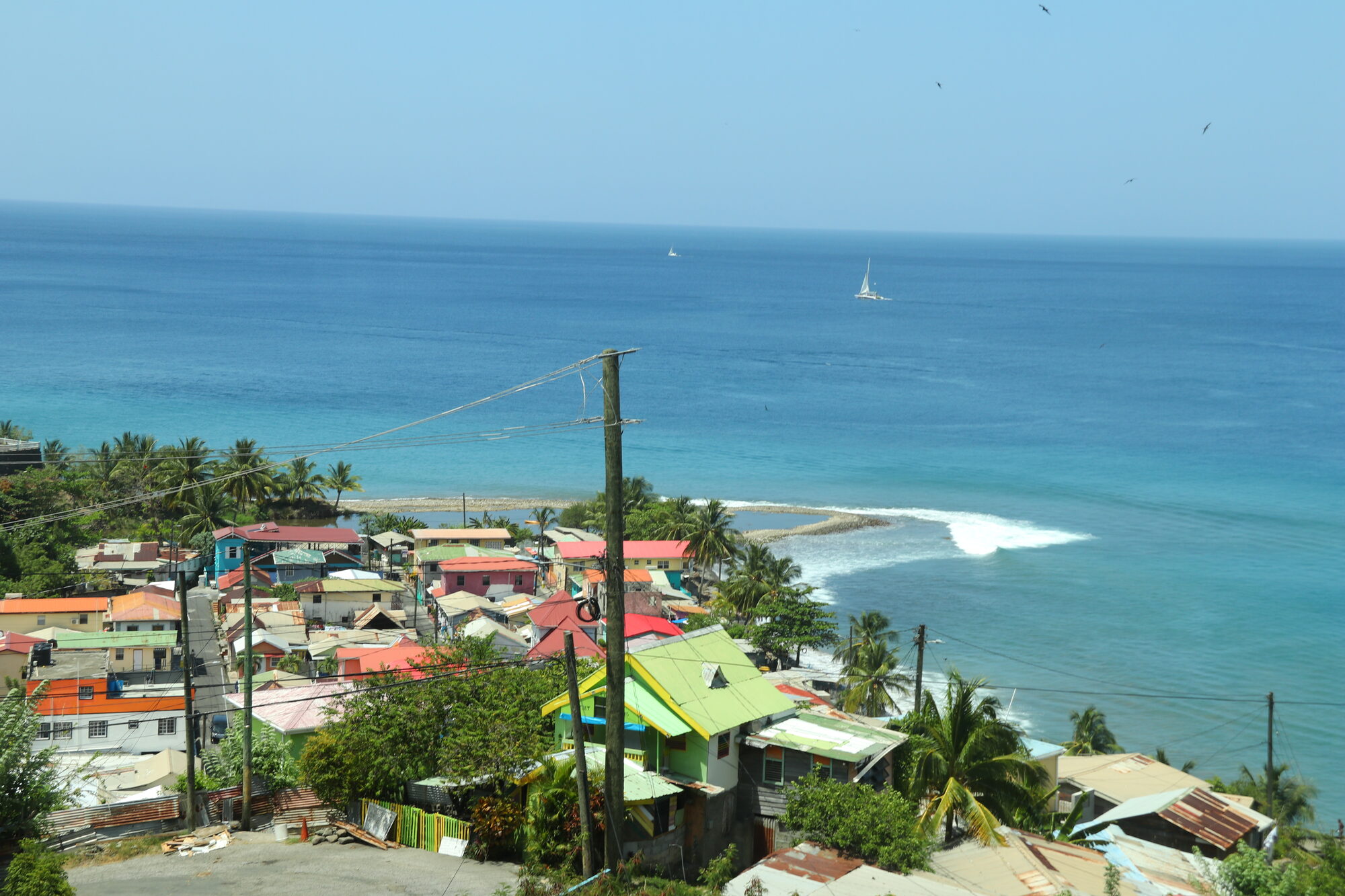 Colorful village rooftops above turquoise Caribbean water with a distant sailboat